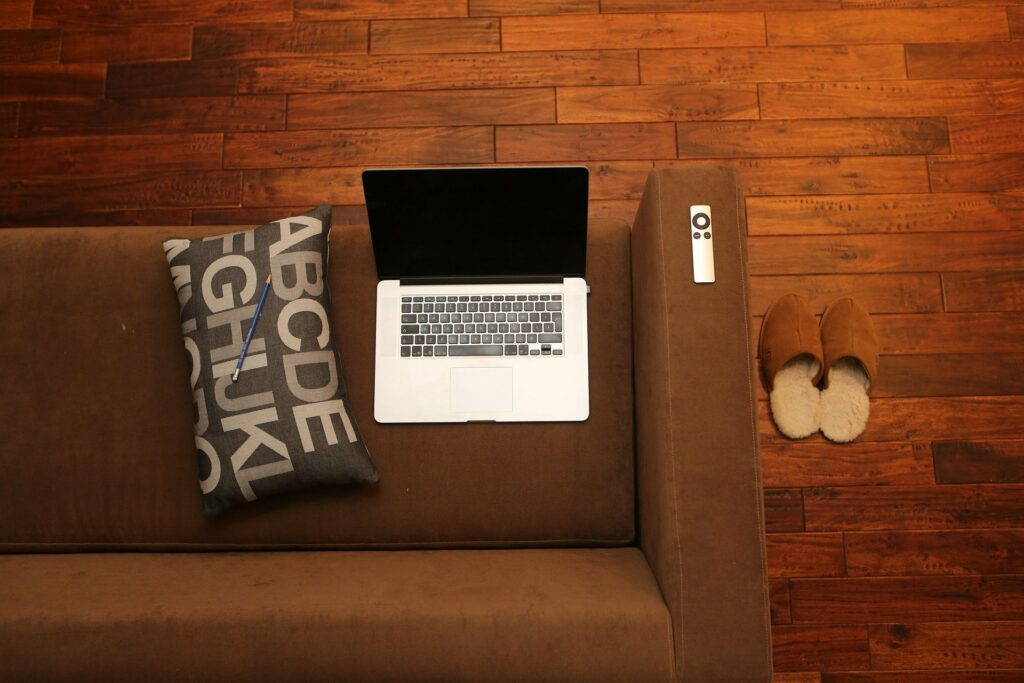 Laptop auf Couch. Neben Laptop ein Kissen auf dem ein Bleistift liegt. Rechts neben der Couch stehen Pantoffeln auf der Erde. Remotearbeit im Home Office